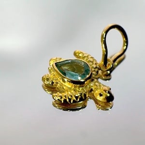 Op de afbeelding: Een goudkleurige schildpendant met een peervormige, teal-kleurige edelsteen. De hanger heeft een lus voor een ketting en is zeer gedetailleerd, met getextureerde oppervlakken en een gepolijste afwerking.