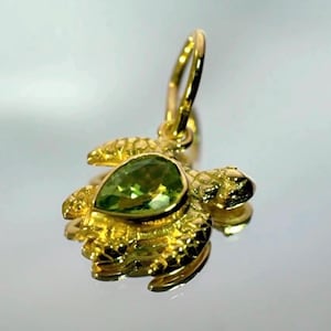 Op de afbeelding: Een goudkleurige schildpadhanger met een peervormige, lichtgroene edelsteen in het midden. De hanger heeft een getextureerd oppervlak en een lus voor een ketting. De kop en vinnen van de schildpad zijn gedetailleerd.