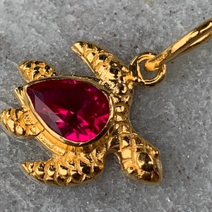 Op de afbeelding: Gouden schildpad hanger met een rode edelsteen. De schildpad heeft een gedetailleerde schild en een lus om aan een ketting te bevestigen.