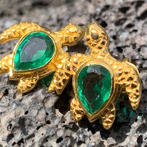 May include: A pair of gold turtle earrings with green gemstone accents. The turtles are detailed with textured shells and have a realistic design.