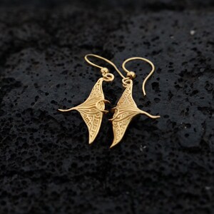 May include: A pair of gold-toned earrings shaped like manta rays. The earrings are detailed with intricate patterns and hang from small loops.