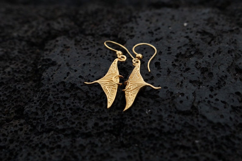 May include: A pair of gold-toned earrings shaped like manta rays. The earrings are detailed with intricate patterns and hang from small loops.