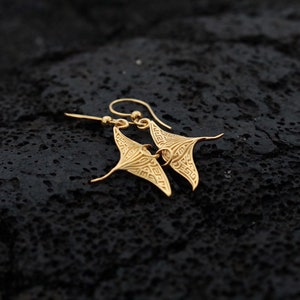 May include: Gold-toned earrings shaped like manta rays with intricate designs. The earrings are hanging from a black surface.