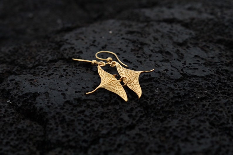 May include: Gold-toned earrings shaped like manta rays with intricate designs. The earrings are hanging from a black surface.