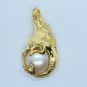 Op de afbeelding: Een gouden zeemeermin hanger met een grote, iriserende parel. De zeemeermin is om de parel gekruld, met haar staart als basis van de hanger.