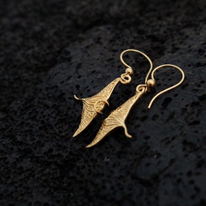 May include: A pair of gold-toned earrings with a manta ray design. The earrings are delicate and have a dangling style.