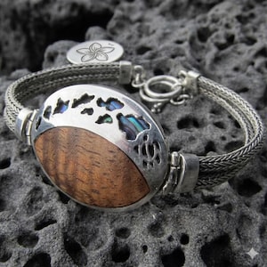 May include: A silver bracelet featuring a woven chain and a central oval focal point. The focal point combines a smooth, brown wood inlay with a silver overlay displaying cut-out shapes filled with iridescent abalone shell and a flower charm.