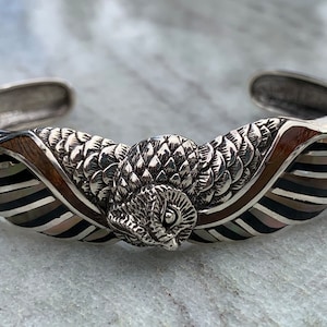 May include: Silver cuff bracelet featuring an owl design. The bracelet has a detailed owl centerpiece with textured feathers. The band incorporates wood and iridescent stripes in shades of black, gray, and brown, creating a unique and artistic accessory.