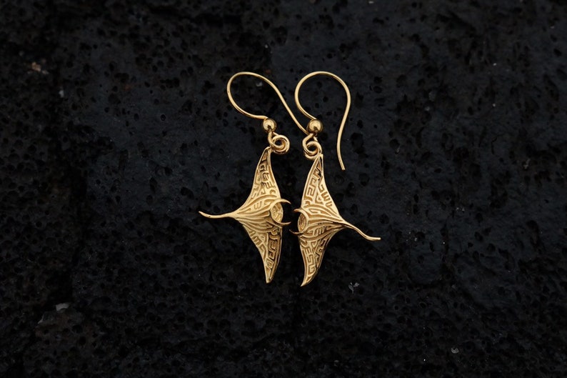 May include: A pair of gold-tone earrings featuring a detailed manta ray design. The earrings are made of metal and have a dangling style.