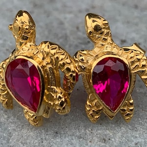 May include: A pair of gold turtle earrings with pink gemstone accents. The turtles are detailed with textured shells and have a three-dimensional design.