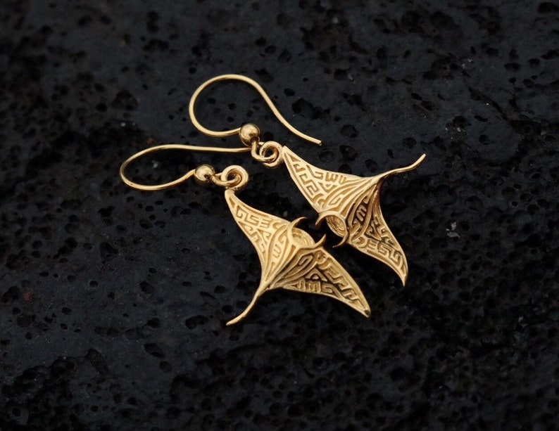 May include: A pair of gold-toned earrings shaped like manta rays with intricate geometric designs. The earrings have a dangling design and are made of metal.