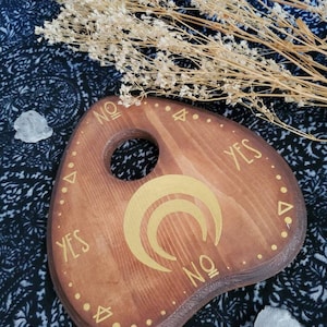 May include: Wooden planchette with a gold crescent moon design and the words "YES", "NO", and "YES" written around the edge.