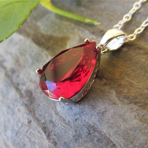 May include: A silver pendant necklace with a large, teardrop-shaped, faceted red gemstone. The gemstone is set in a silver bezel with intricate detailing. The chain is silver, and the necklace is displayed on a textured grey surface.