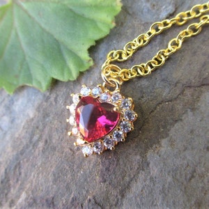 May include: A gold chain necklace with a heart-shaped pendant. The pendant features a large, red gemstone surrounded by smaller, clear gemstones.