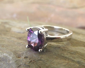 May include: A silver ring with a large, oval-shaped purple gemstone set in a prong setting. The ring is on a gray, textured surface.