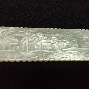 May include: Rectangular, off-white carved mother-of-pearl plaque. The plaque features a detailed relief scene of figures near buildings and trees. The edges are scalloped. The carving is intricate and the overall effect is delicate and ornate.