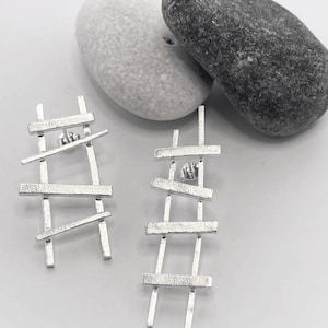 Könnte beinhalten: Ein Paar silberne Ohrringe mit geometrischem Design. Die Ohrringe bestehen aus dünnen, flachen Metallstücken, die in einer leiterartigen Form angeordnet sind. Die Ohrringe sind modern und minimalistisch.