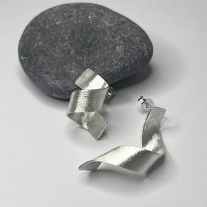 Könnte beinhalten: Ein Paar silberne Ohrringe mit einem gedrehten, bandartigen Design. Die Ohrringe sind aus gebürstetem Silber gefertigt und haben einen modernen, minimalistischen Stil.