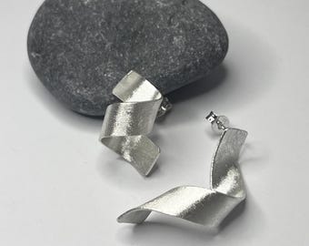 Könnte beinhalten: Ein Paar silberne Ohrringe mit einem gedrehten, bandartigen Design. Die Ohrringe sind aus gebürstetem Silber gefertigt und haben einen modernen, minimalistischen Stil.