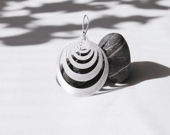 Puede incluir: Pendientes de plata con un diseño circular que presenta múltiples círculos concéntricos. Los pendientes se muestran sobre una superficie blanca con una piedra lisa de color gris.