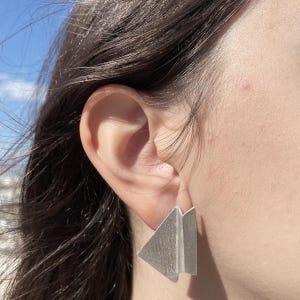May include: A silver geometric earring with a triangular design. The earring is made of a brushed metal material and has a modern, minimalist style. The earring is worn on the ear.