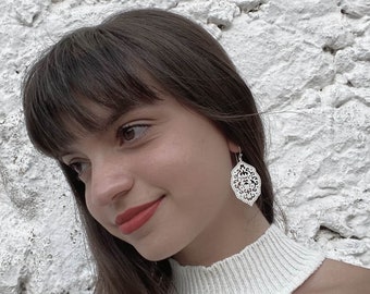 Puede incluir: Una mujer con cabello castaño lleva un suéter blanco y pendientes de filigrana plateada. Los pendientes tienen forma de hoja con un diseño delicado e intrincado.