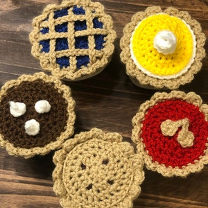 May include: Five crocheted pie decorations in various colors and designs. The pies are brown, blue, yellow, red, and tan.