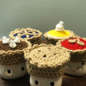 May include: Four crocheted pie mug toppers in different flavors: chocolate, blueberry, pumpkin, and cherry. The toppers are on top of white mugs with a smiling face.