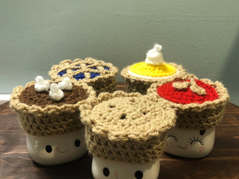 May include: Four crocheted pie mug toppers in different flavors: chocolate, blueberry, pumpkin, and cherry. The toppers are on top of white mugs with a smiling face.