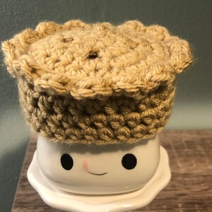 May include: A crocheted pie crust hat in a beige color sits on top of a white ceramic mug with a smiling face.