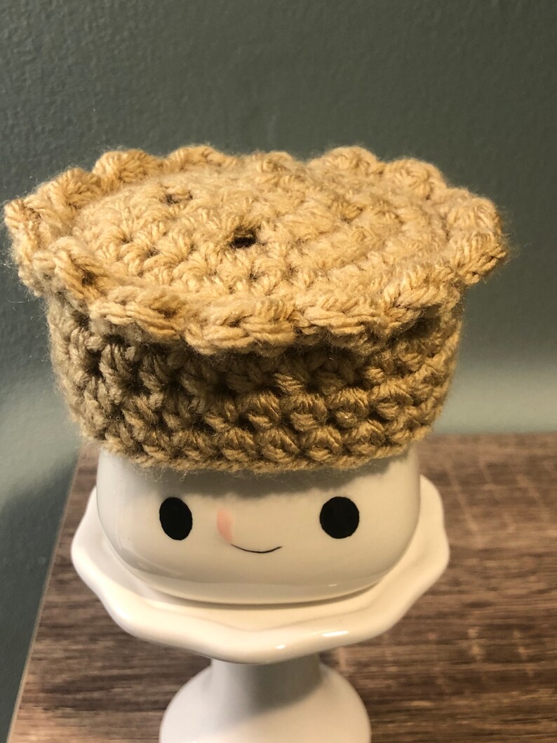 May include: A crocheted pie crust hat in a beige color sits on top of a white ceramic mug with a smiling face.