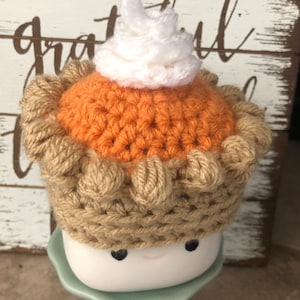 May include: A crocheted pumpkin pie mug cozy with a white whipped cream topping. The cozy is brown and orange and has a cute face with black eyes. The cozy is sitting on a light green pedestal.