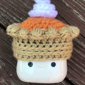 May include: A white ceramic mug with a crocheted pumpkin pie hat. The hat has a brown crust, an orange filling, and a white whipped cream topping.