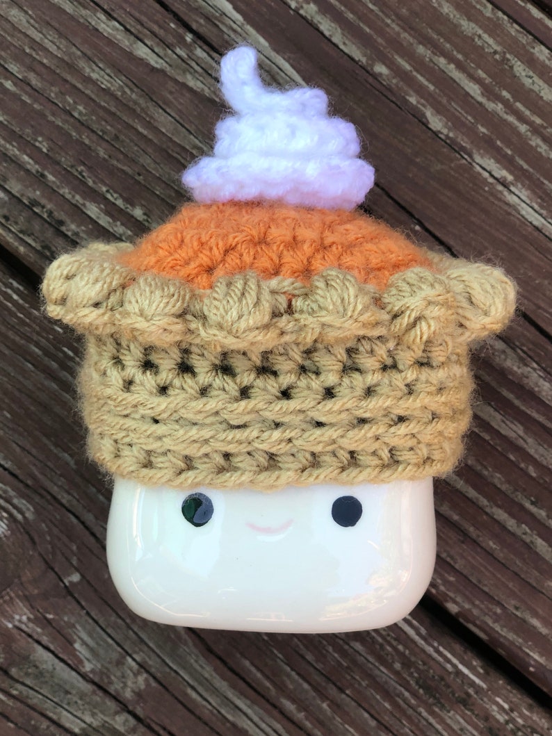 May include: A white ceramic mug with a crocheted pumpkin pie hat. The hat has a brown crust, an orange filling, and a white whipped cream topping.