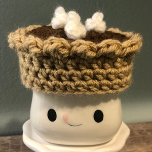 May include: A white ceramic mug with a smiling face, wearing a brown crocheted pie crust hat with white fluffy marshmallows on top.