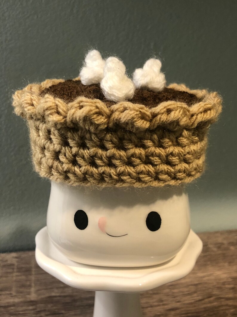 May include: A white ceramic mug with a smiling face, wearing a brown crocheted pie crust hat with white fluffy marshmallows on top.