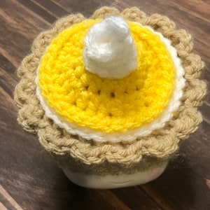 May include: A crocheted pumpkin pie mug cozy with a yellow pie crust, white whipped cream, and a brown rim.