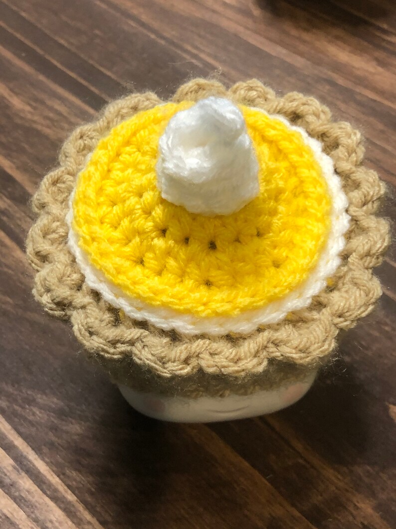 May include: A crocheted pumpkin pie mug cozy with a yellow pie crust, white whipped cream, and a brown rim.