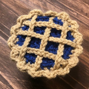 May include: A crocheted pie with a blue filling and a tan crust. The pie is made with yarn and has a woven pattern.