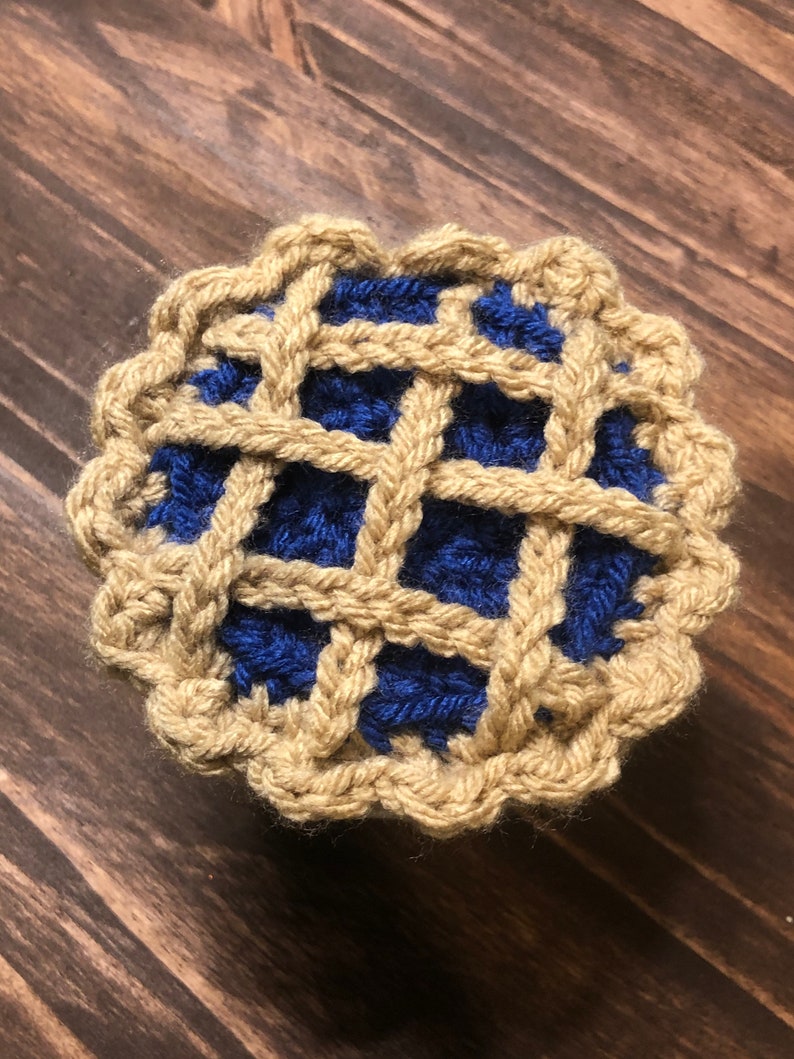 May include: A crocheted pie with a blue filling and a tan crust. The pie is made with yarn and has a woven pattern.