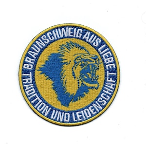 Könnte beinhalten: Ein runder, blauer und gelber gestickter Aufnäher mit einem Löwenkopf in der Mitte. Der Text um den Löwen lautet "Tradition und Leidenschaft aus Braunschweig".