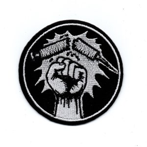 Op de afbeelding: Zwart-witte geborduurde patch met een vuist die twee gebroken spuiten vasthoudt. De vuist wordt omringd door een zwarte cirkel met een witte ster.