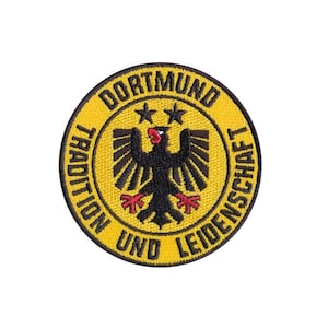 Könnte beinhalten: Ein gelber runder Aufnäher mit schwarzem Text, der "Dortmund Tradition und Leidenschaft" lautet, und einem schwarzen Adler mit roten Akzenten in der Mitte.