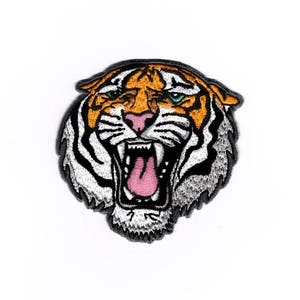 Può includere: Un toppa ricamata con la testa di una tigre. La tigre ha strisce arancioni e nere, un viso bianco e una lingua rosa. Gli occhi della tigre sono blu.
