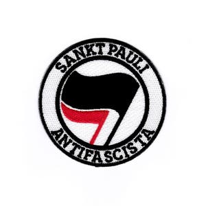 Puede incluir: Un parche circular blanco con texto negro que dice "Sankt Pauli Antifa", un símbolo de puño estilizado rojo y negro, y una bandera negra.