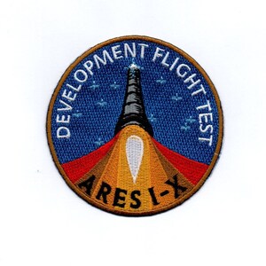 Peut inclure: Un écusson rond brodé de couleur bleu, blanc, rouge et orange avec le texte "Development Flight Test" et "Ares I-X".