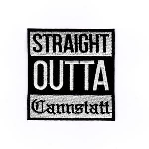 以下が含まれることがあります： 「STRAIGHT OUTTA」と「Cannstatt」の文字が刺繍された白黒のパッチ。 パッチは長方形で、黒い背景に白い文字が配置されています。