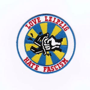 Könnte beinhalten: Ein runder, weißer Patch mit einem blau-gelb gestreiften Hintergrund. Der Patch trägt den Text "Love Leipzig Hate Fascism" in roten Buchstaben am Rand. In der Mitte des Patches befindet sich eine Schwarzweißillustration von zwei ineinander verschlungenen Händen.