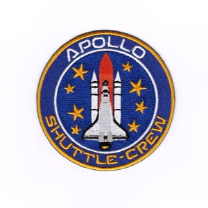May include: Circular embroidered patch with the Apollo Shuttle Crew logo. The patch has a blue background with gold stars. The text "APOLLO" and "SHUTTLE-CREW" are in white. A rocket ship is in the centre, with red, white, and grey details.
