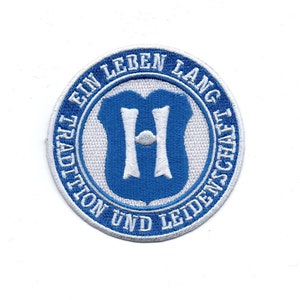 Könnte beinhalten: Ein blauer und weißer gestickter Aufnäher mit dem Text "TRADITION UND LEIDENSCHAFT EIN LEBEN LANG" in einem Kreis um einen blauen Schild mit einem weißen "H" darin.